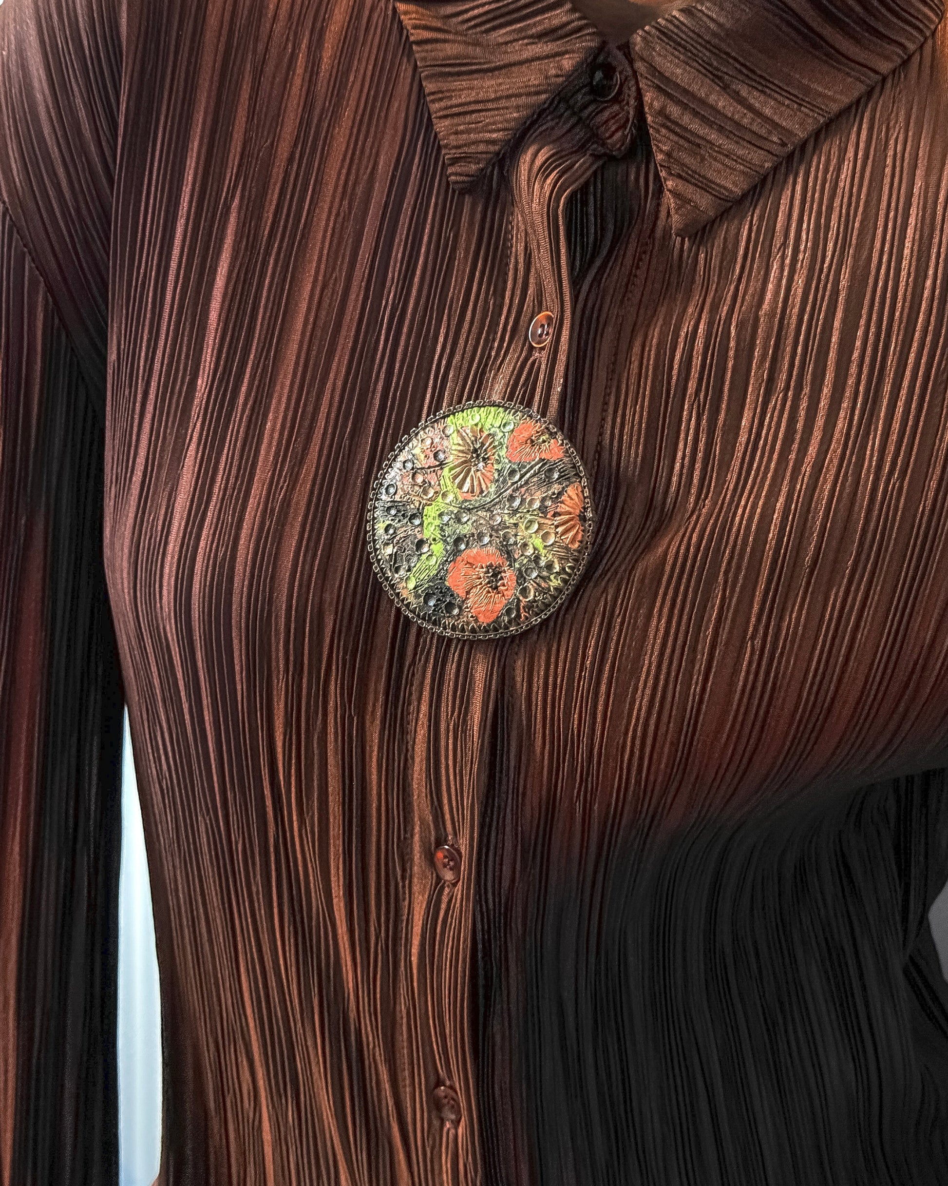 Close-up of a brown pleated shirt with a decorative pendant on a plain background